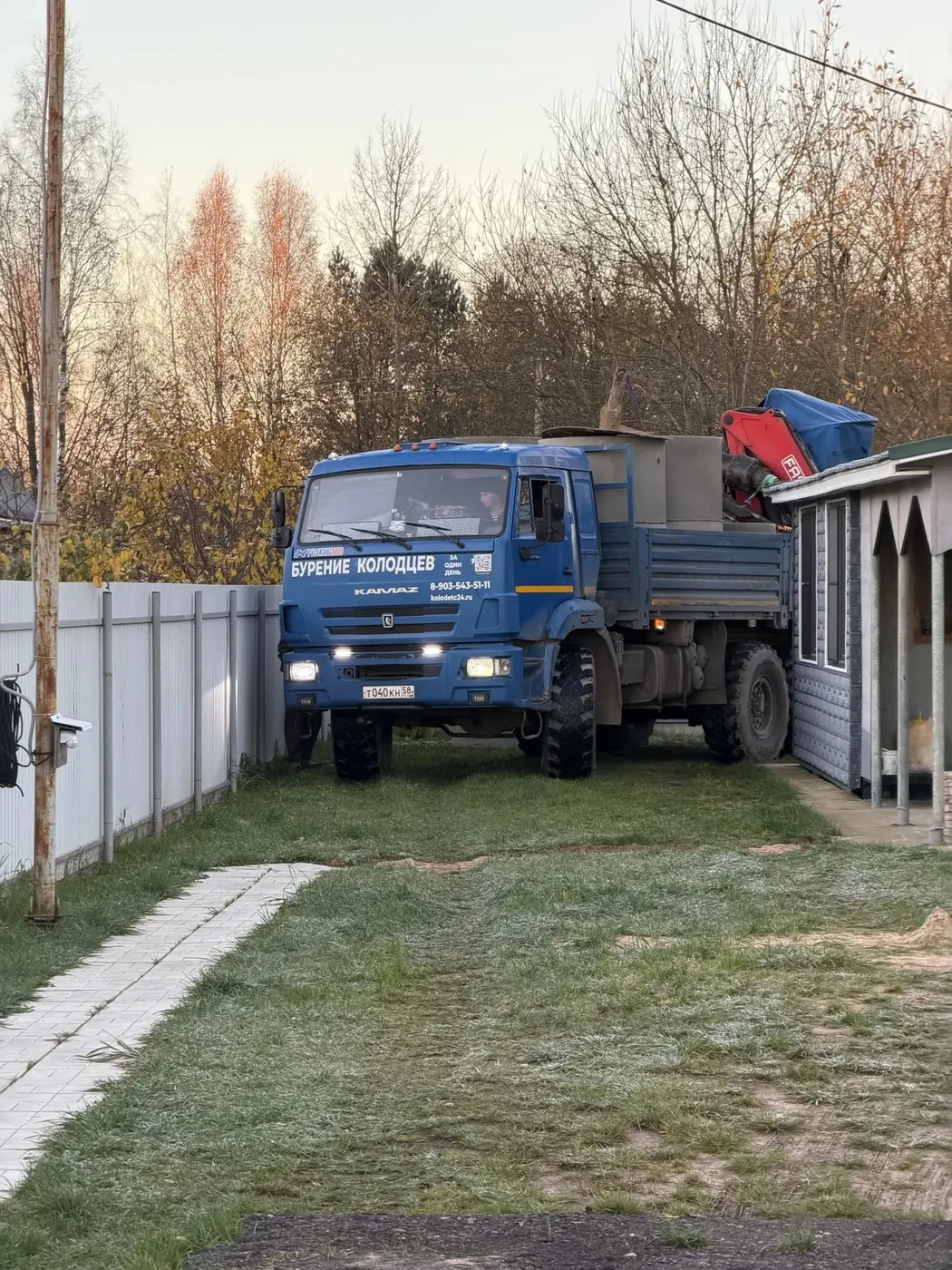 Синий КамАЗ с краном-манипулятором и надписью «Бурение колодцев» на участке у забора